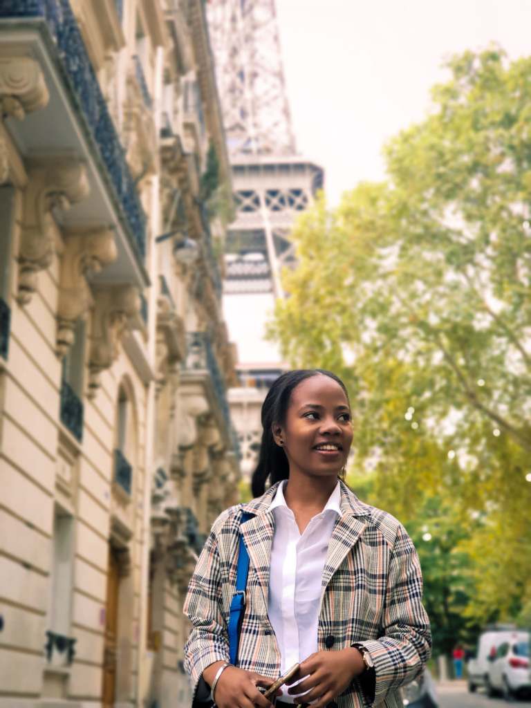 Jeune femme urbaine dans une ruelle dans paris, avec la Tour Eiffel en arrière-plan. Elle porte une chemise et une veste à carreaux. Regard vers la droite. Tiens son smartphone